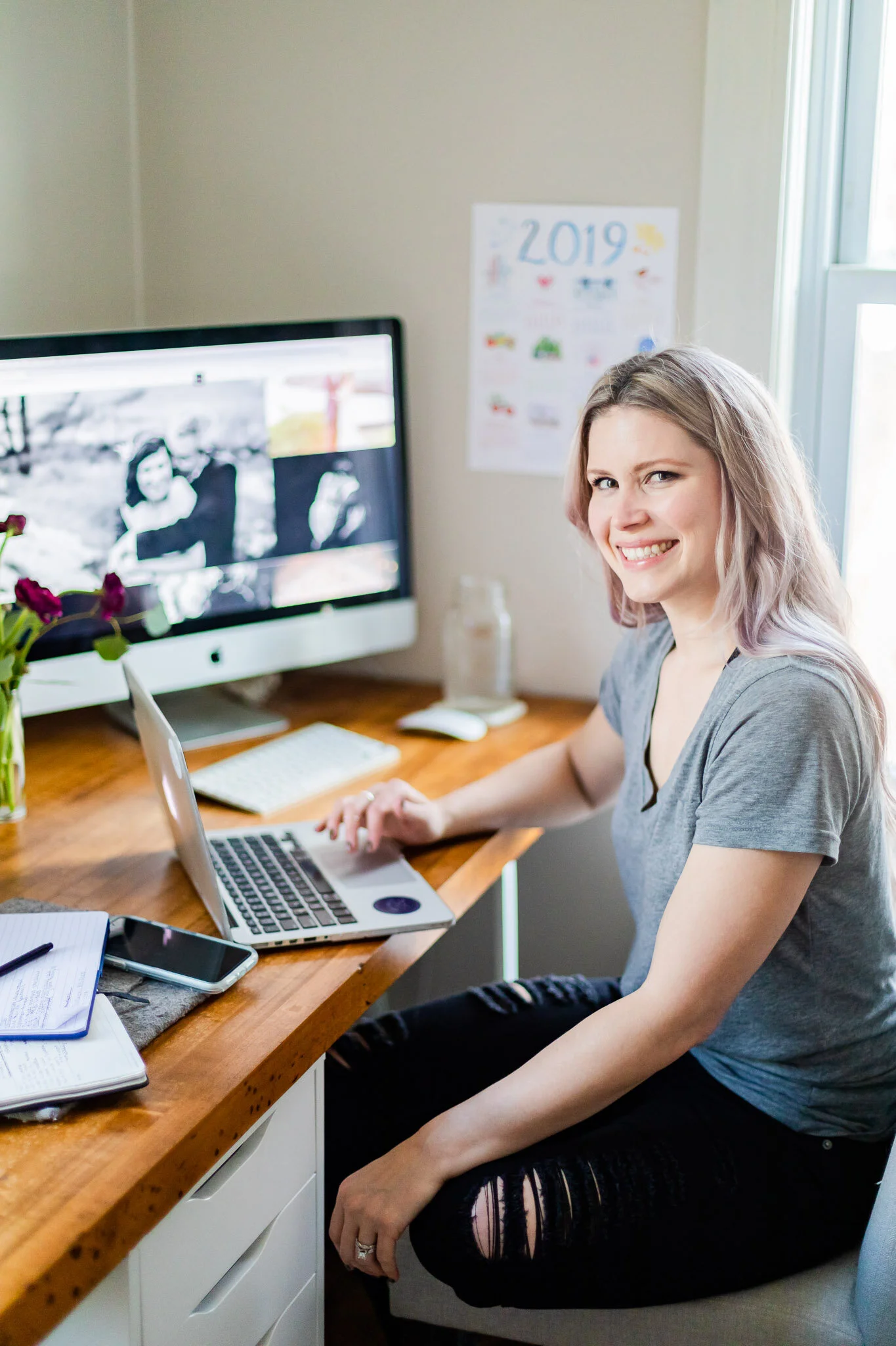 A woman with long hair sitting at a wooden desk using a laptop. She is smiling and an iMac displaying black and white photos is in the background. There are flowers and a notebook on the desk, and a calendar is on the wall.