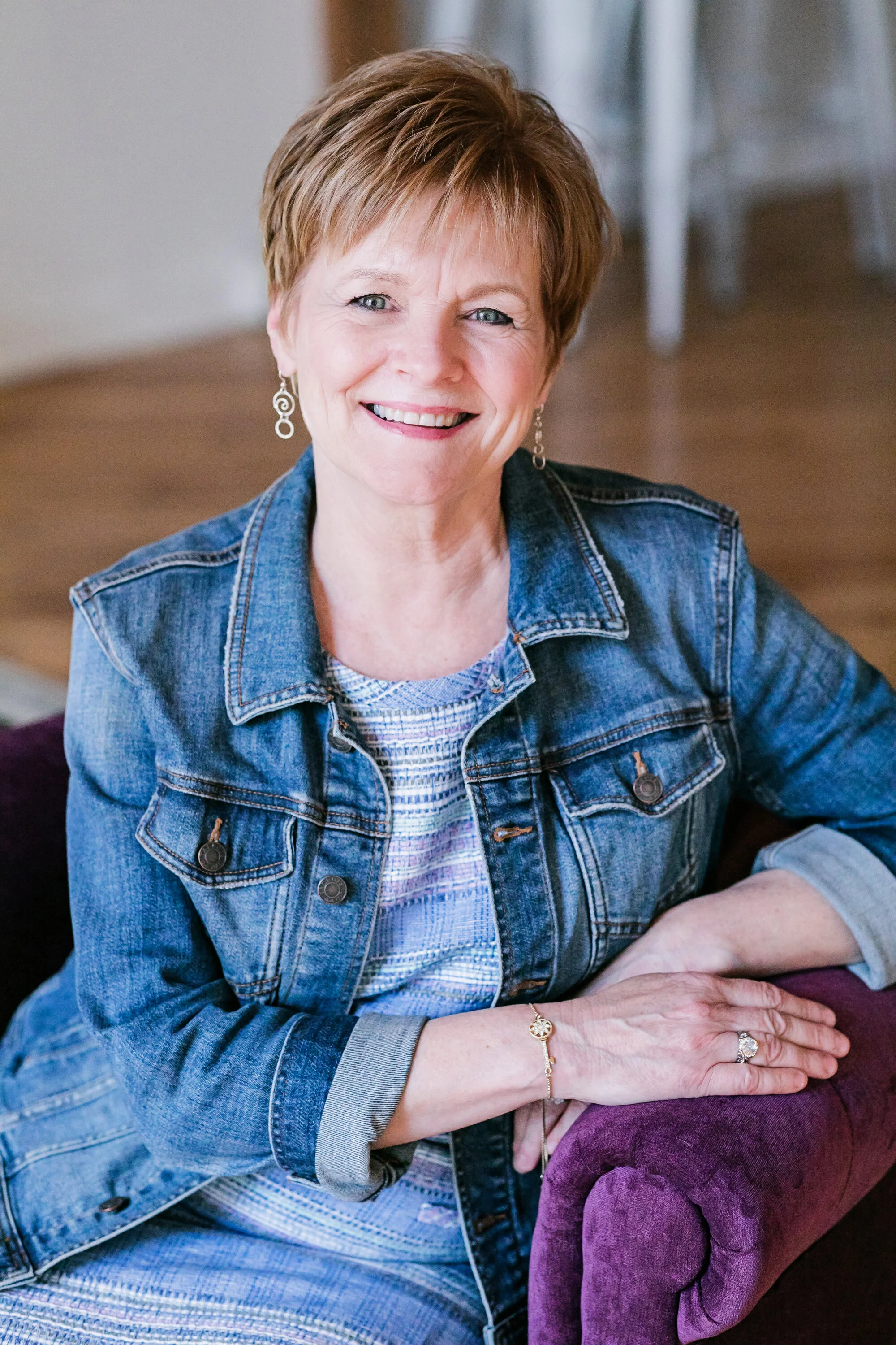 A smiling woman with short reddish hair, wearing a denim jacket with a patterned top underneath, sitting on a purple armchair.