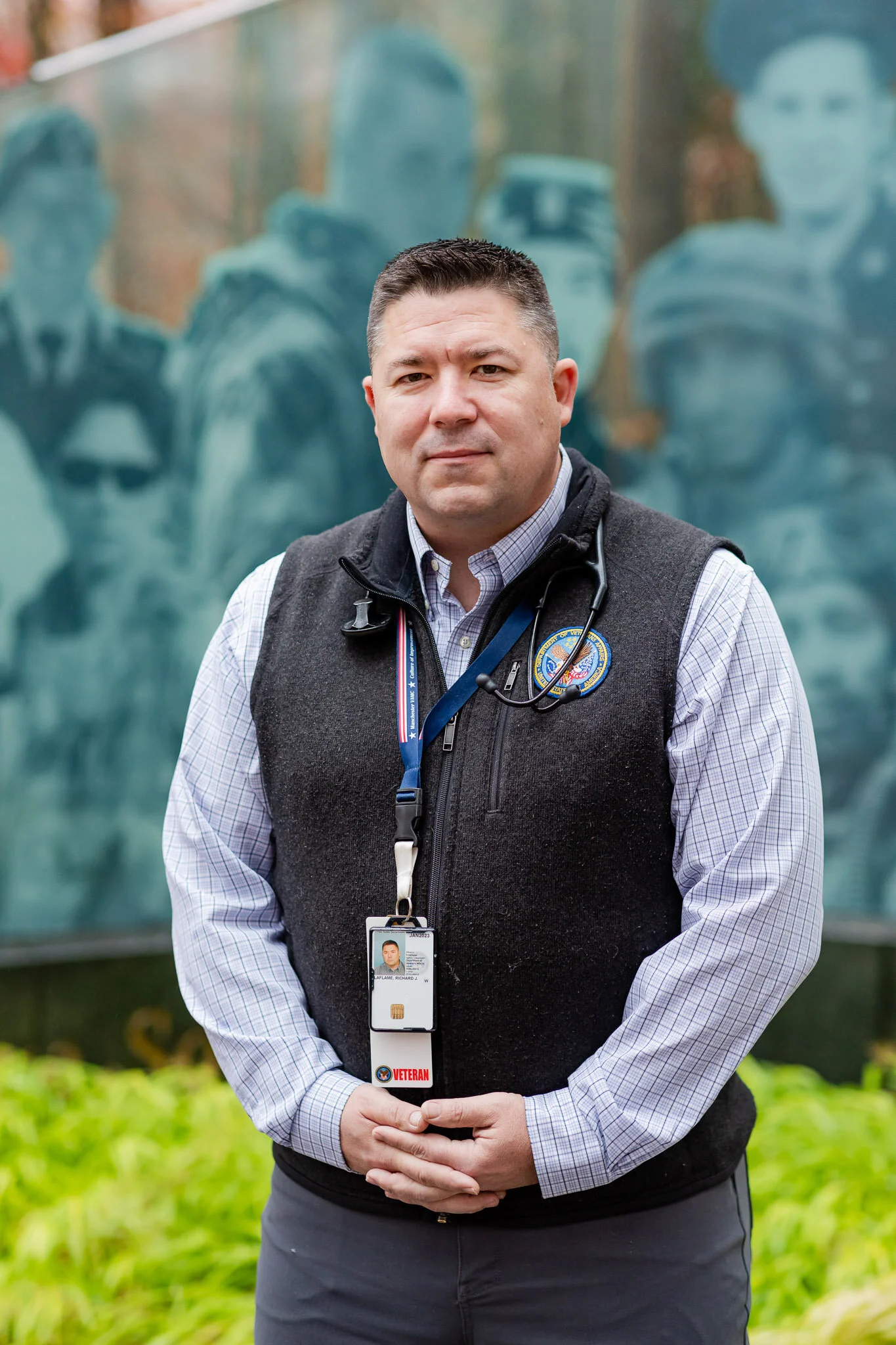 A man wearing a gray vest with a veteran badge and stethoscope stands outdoors in front of a memorial wall with engraved portraits of soldiers.