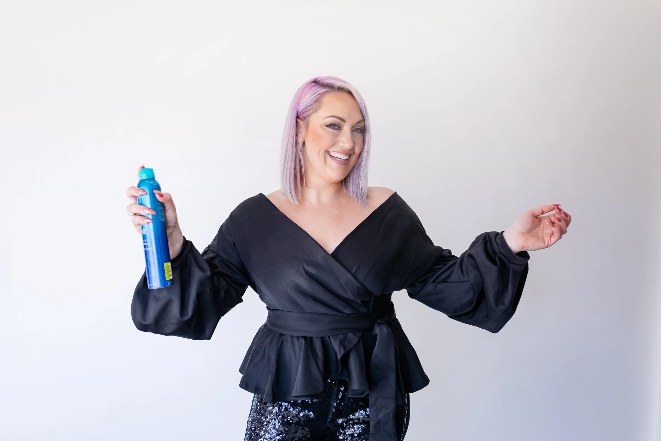 A smiling woman with light purple hair holding a can of aerosol spray in her right hand, wearing a black satin top with puff sleeves and sequined pants, standing against a plain white wall.