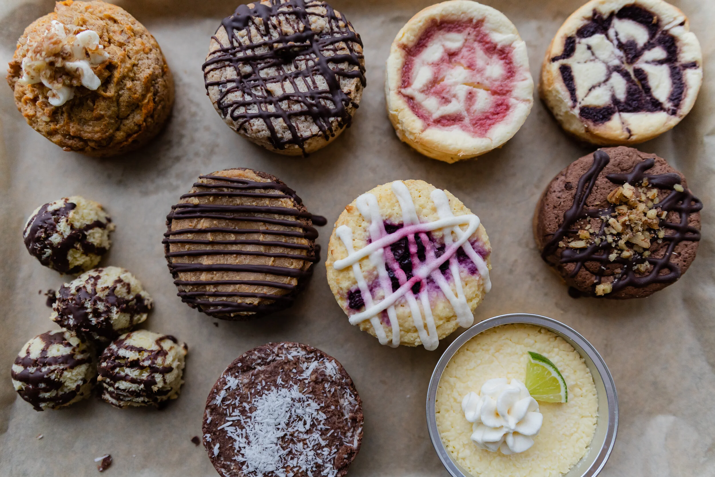Various decorated cupcakes and cookies on parchment paper, with a small container of lime-flavored cheese and whipped cream topping.
