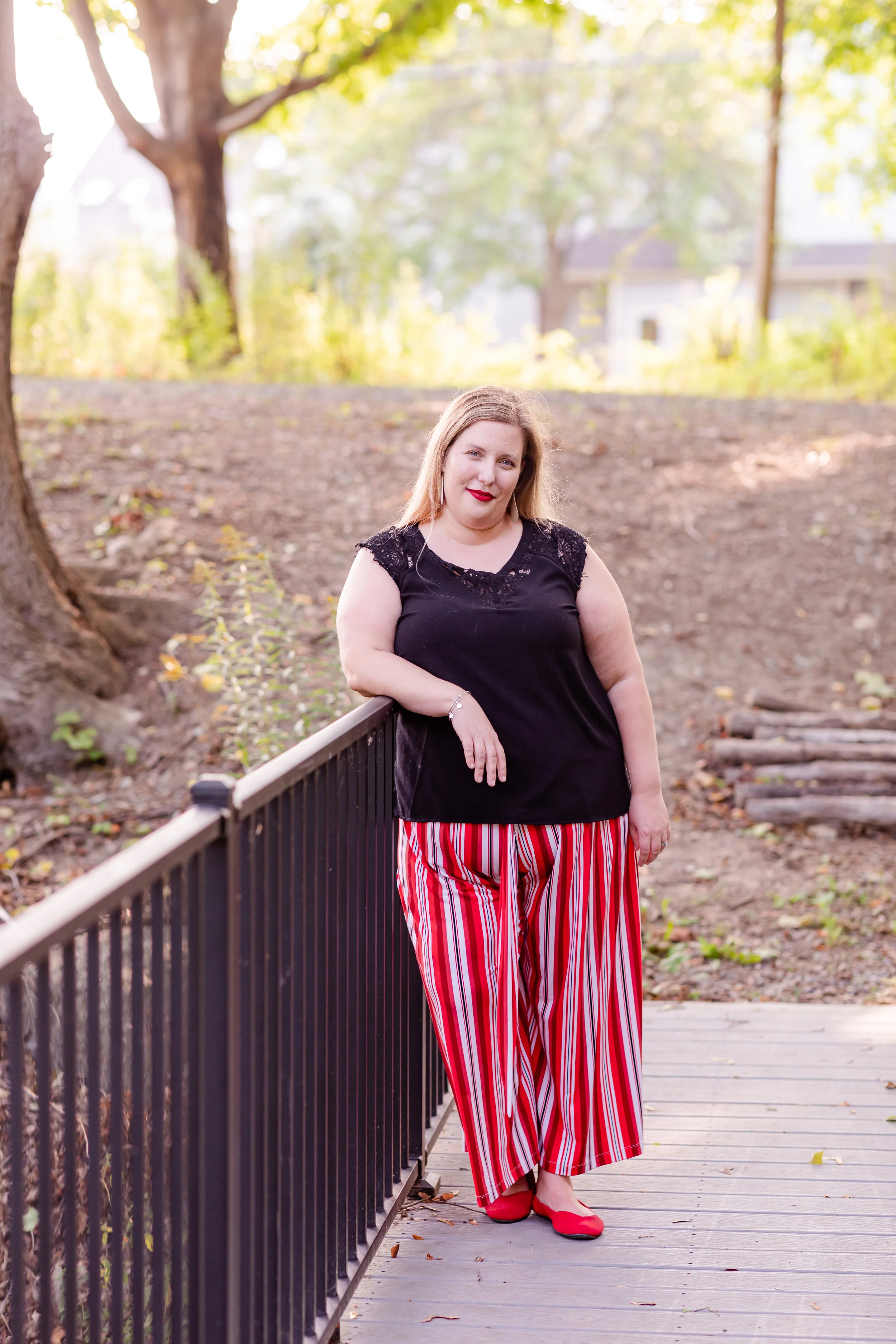 A woman in a black top and red striped pants leaning on a railing outdoors, with trees and houses in the background.