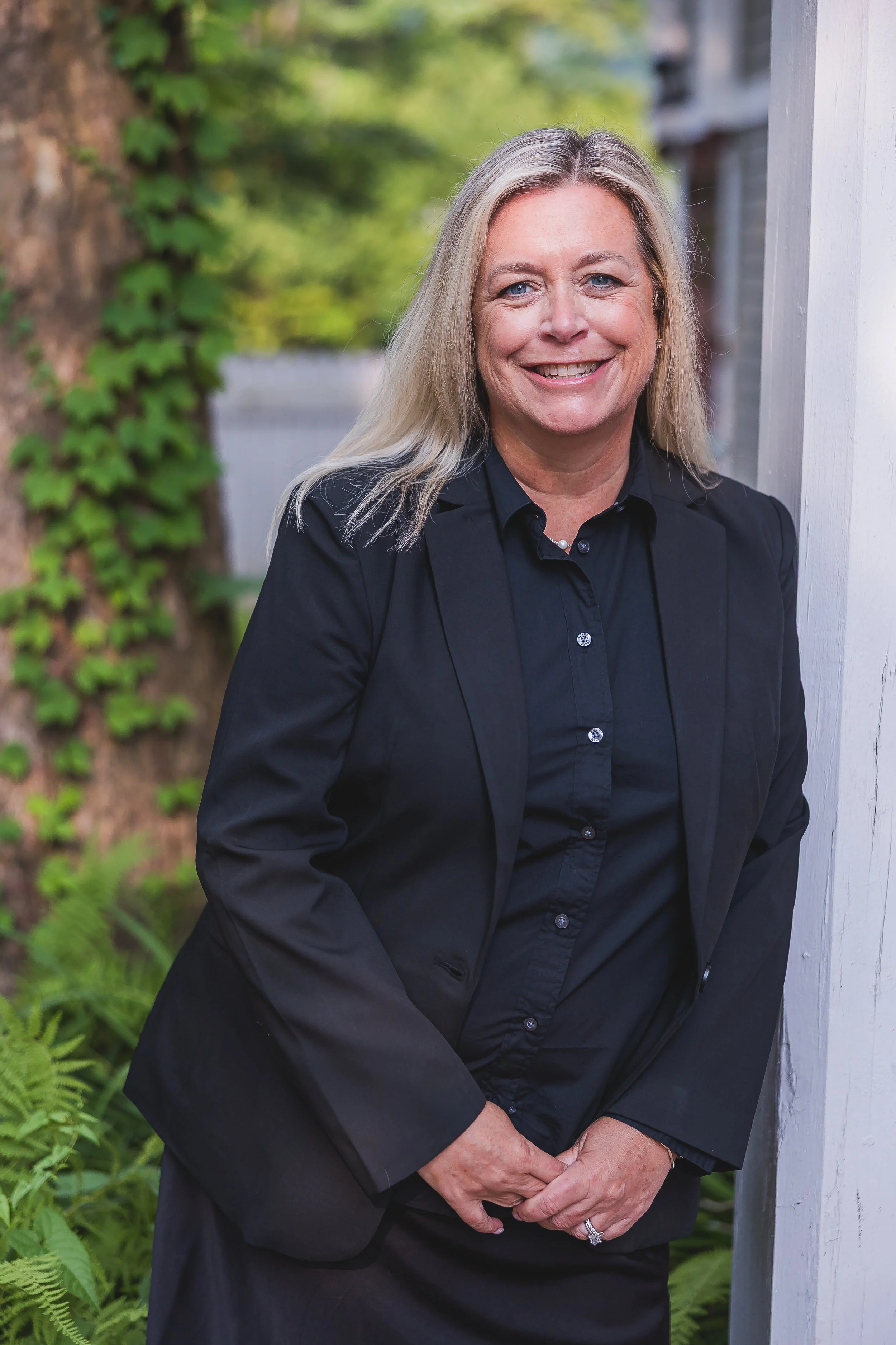 A middle-aged woman with blonde hair wearing a black blazer and black shirt, standing outdoors next to a white wall, smiling.