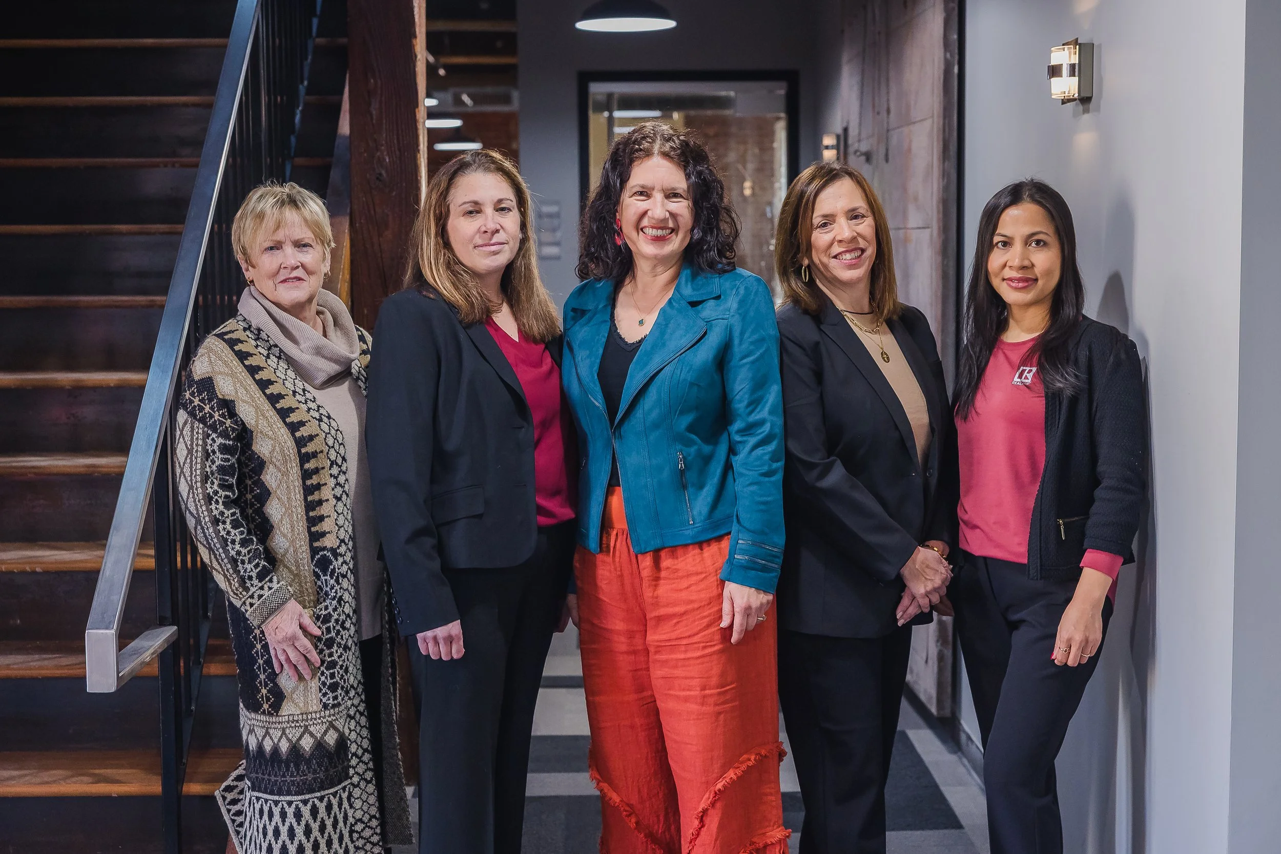 Five women standing together indoors, smiling for the camera near a staircase with a modern interior background.