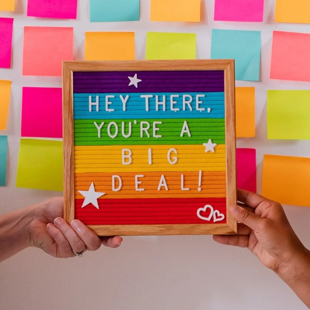 Colorful letter board with message "Hey there, you're a big deal!" held by hands, surrounded by colorful sticky notes on wall.