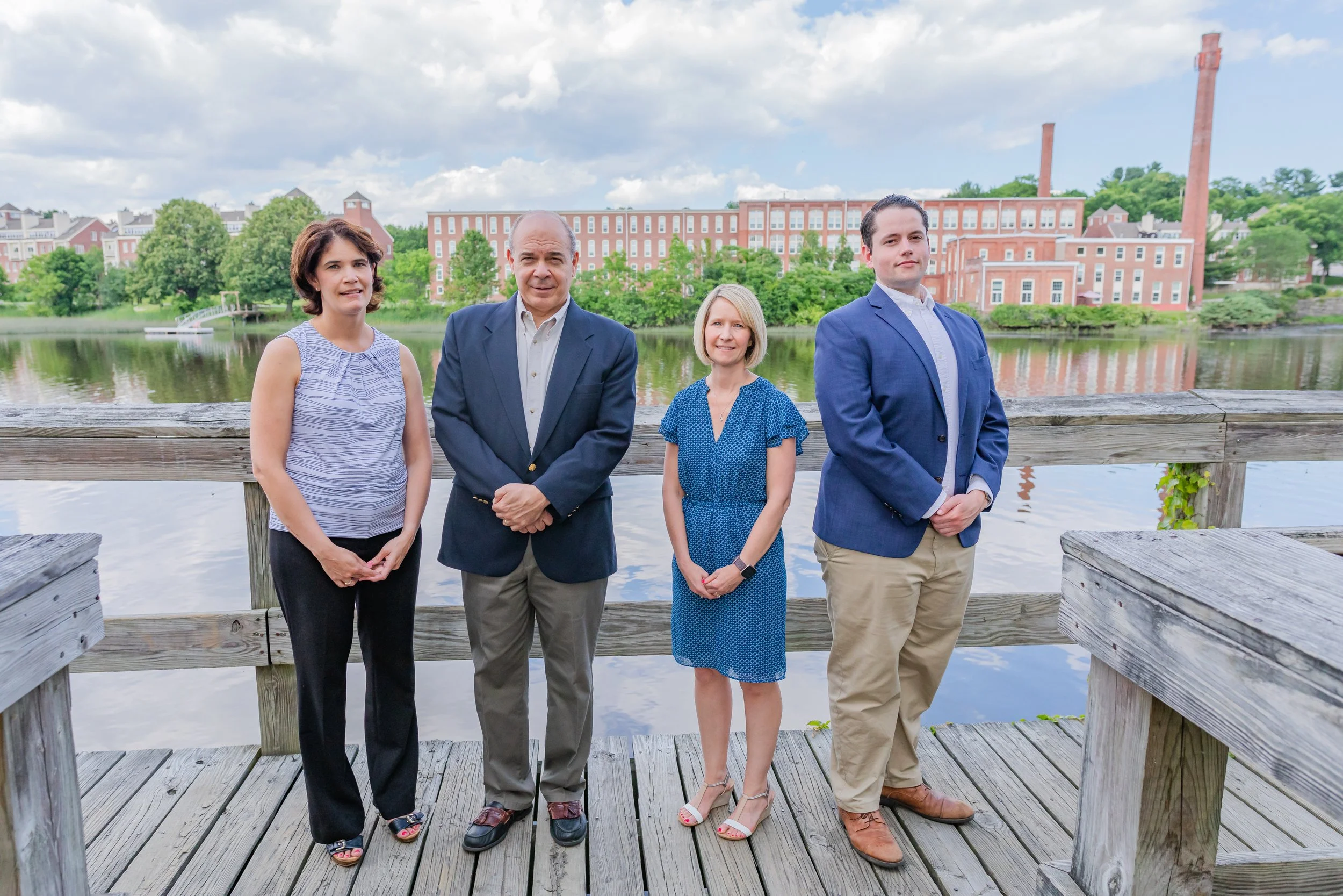 Four people standing on a wooden dock by a lake with a brick building and trees in the background.