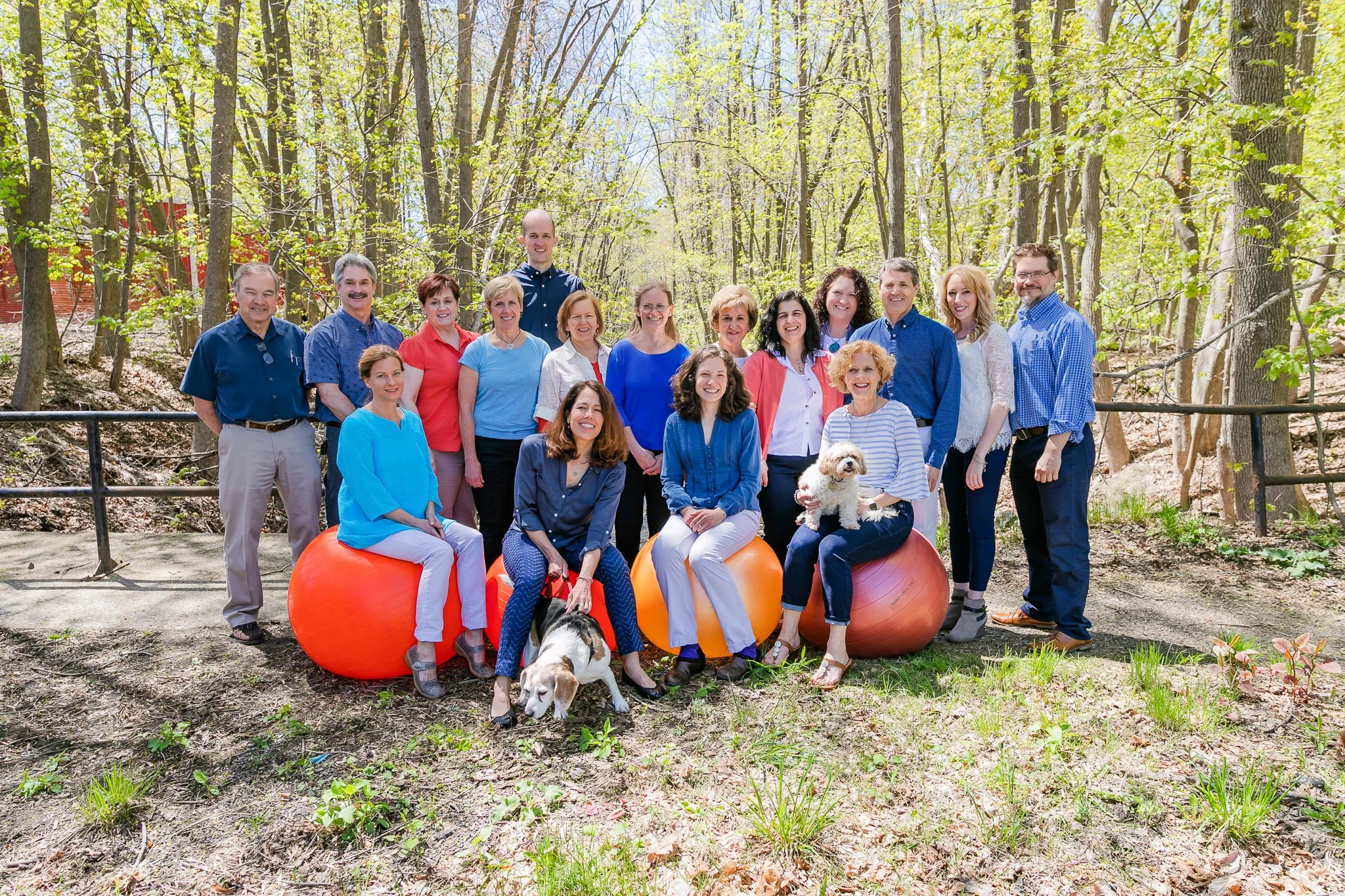 A group of 16 people, including two dogs, standing and sitting on large orange exercise balls outdoors in a wooded park on a sunny day.
