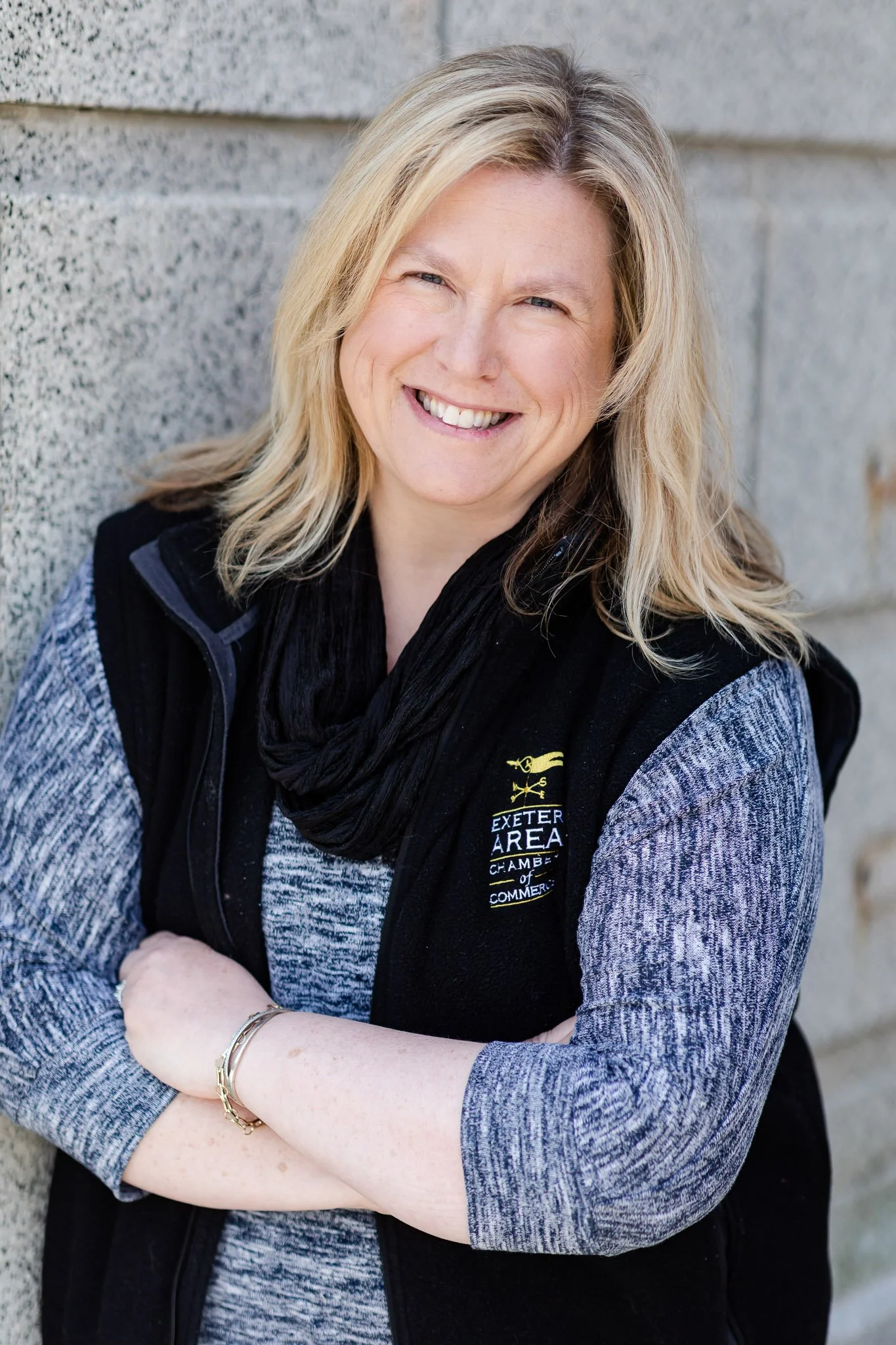A woman with blonde hair smiling, wearing a black vest with an Exeter Area Chamber of Commerce logo, standing against a stone wall with arms crossed.