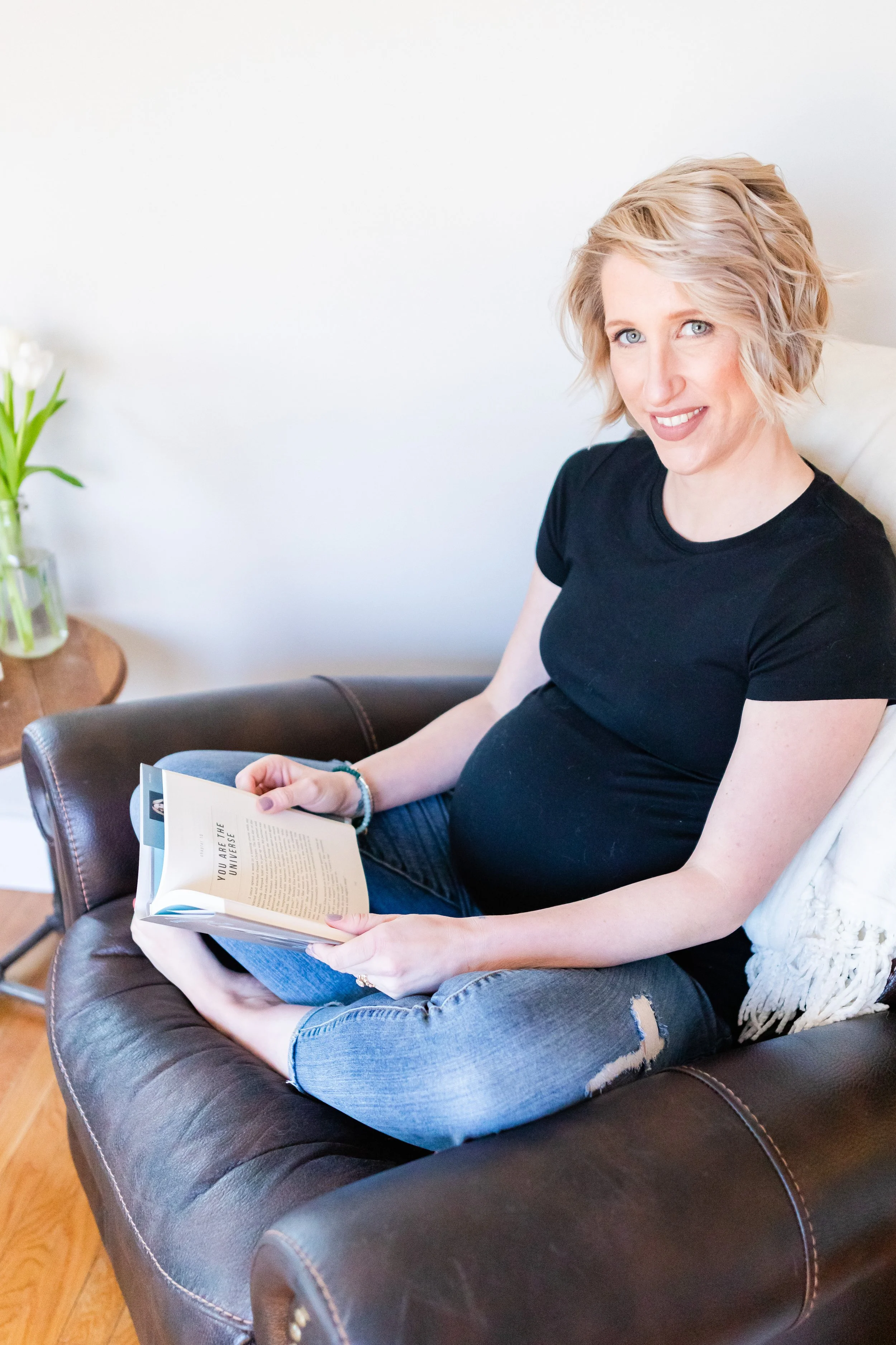 A pregnant woman sitting on a leather couch, reading a book, with a white pillow behind her, a small wooden side table with a glass vase of white flowers, and a white wall in the background.