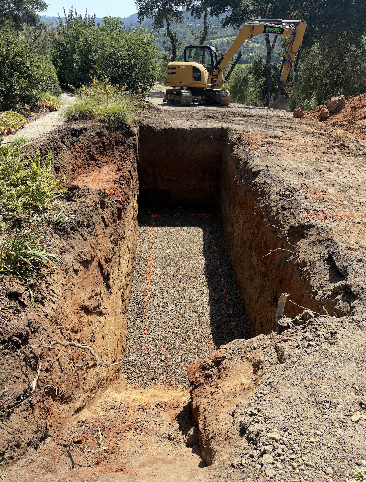 An excavated trench with gravel base, marked with orange spray paint, and a small excavator on the dirt surface at a construction site surrounded by trees and hills.