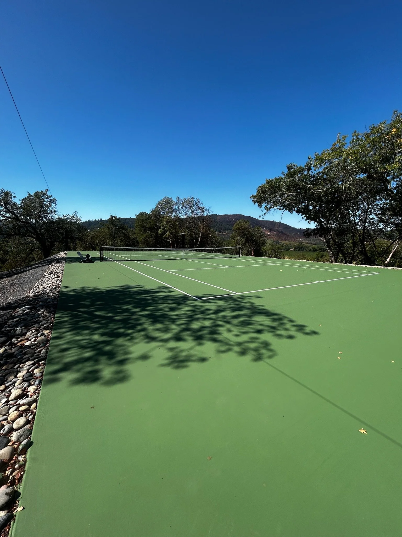 An empty outdoor tennis court with a green surface, surrounded by trees and mountains in the background, under a clear blue sky.