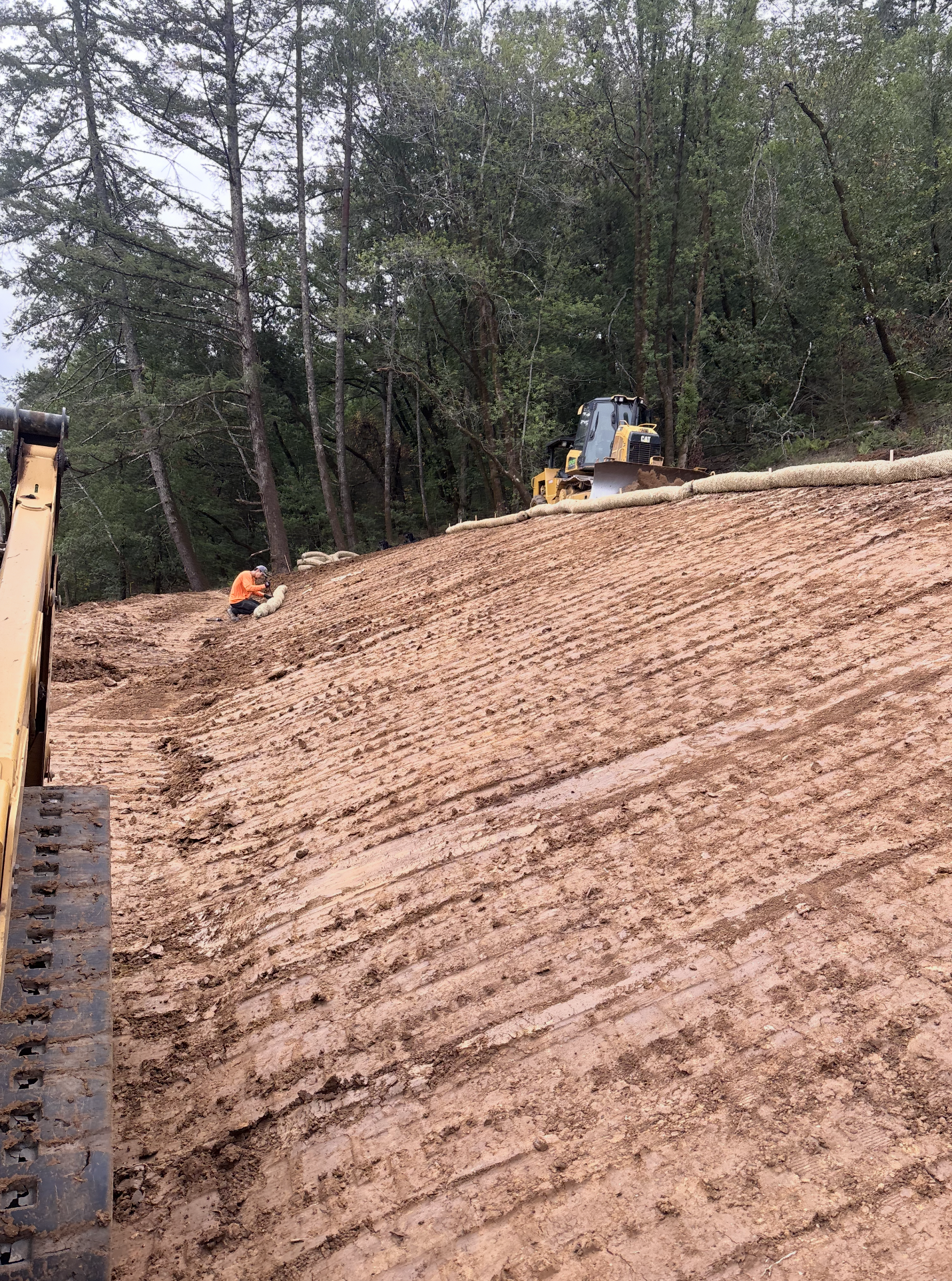 Construction workers leveling a sloped dirt hill with heavy machinery in a wooded area.