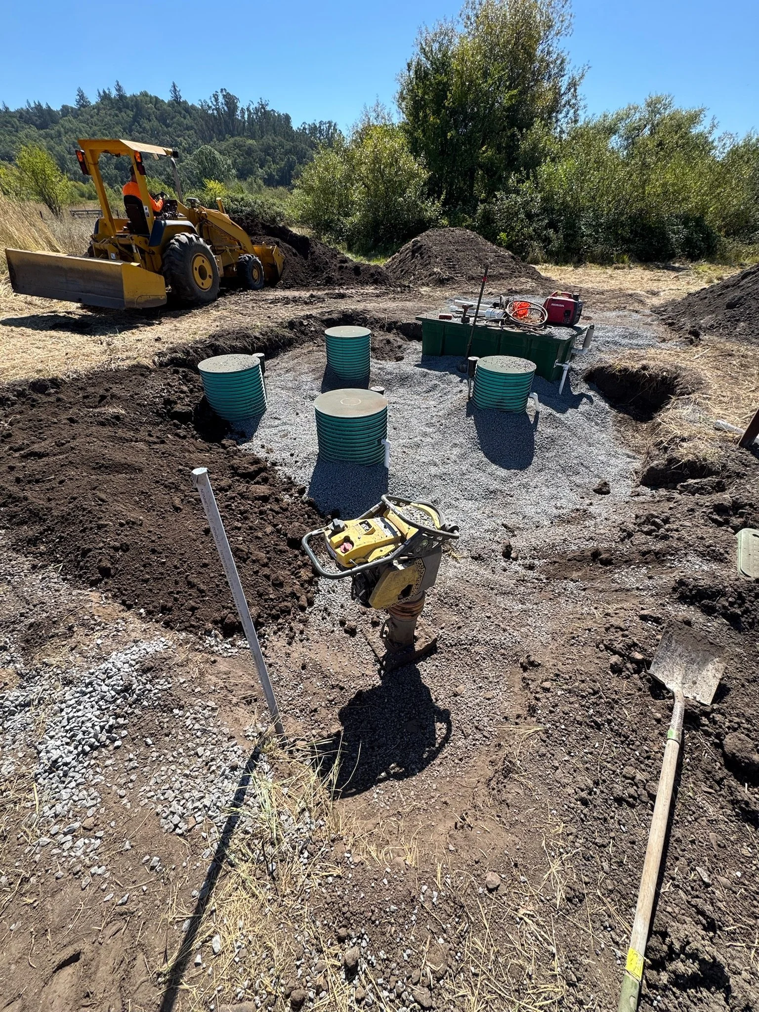 Construction site with a yellow backhoe and men digging trenches, with several green and white pipes laid out on gravel, surrounded by dirt and tools, under clear blue sky.