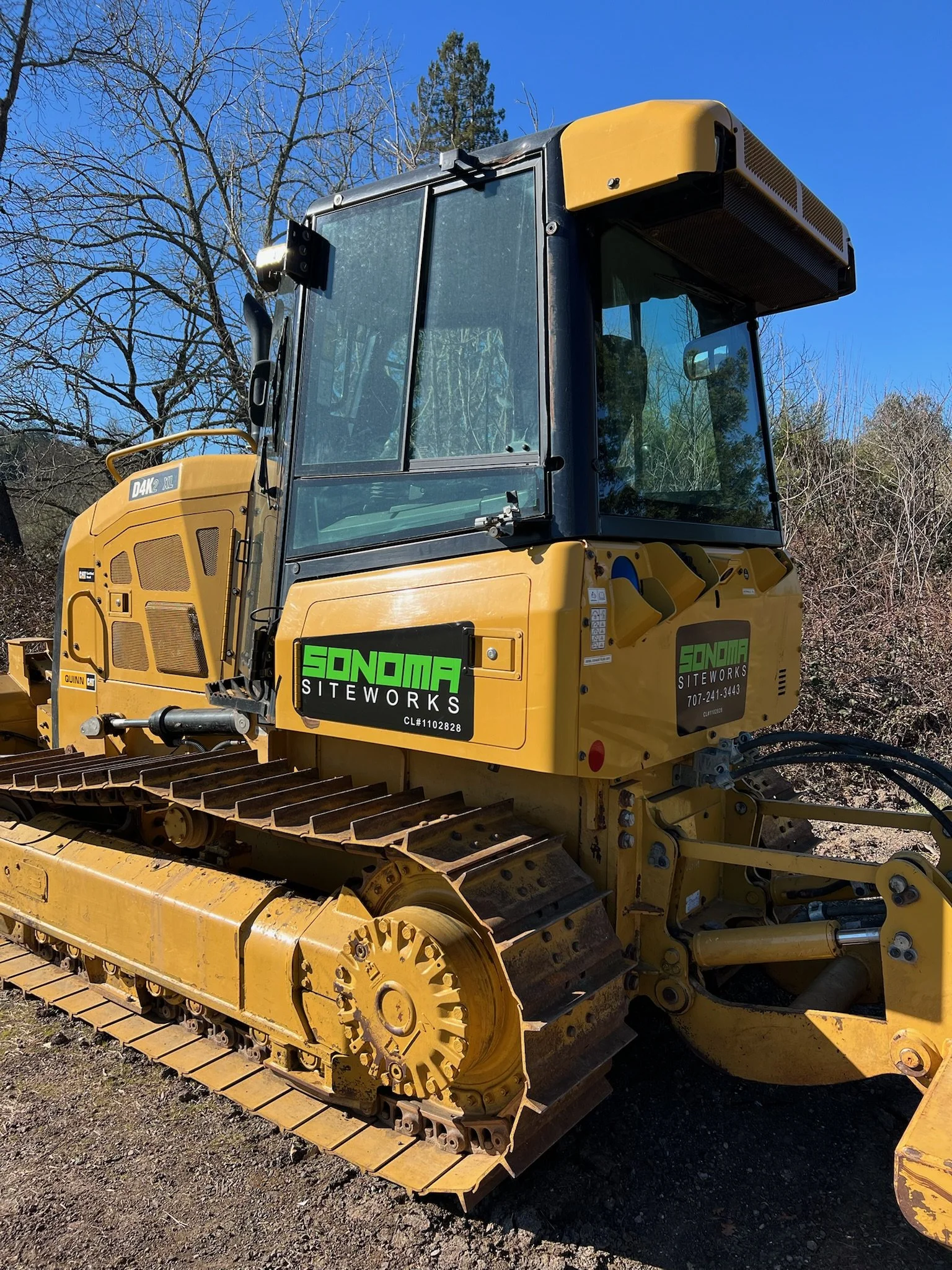 Yellow Caterpillar bulldozer with green and black Sonoma Siteworks stickers, parked outdoors on dirt, with leafless trees and a blue sky in the background.