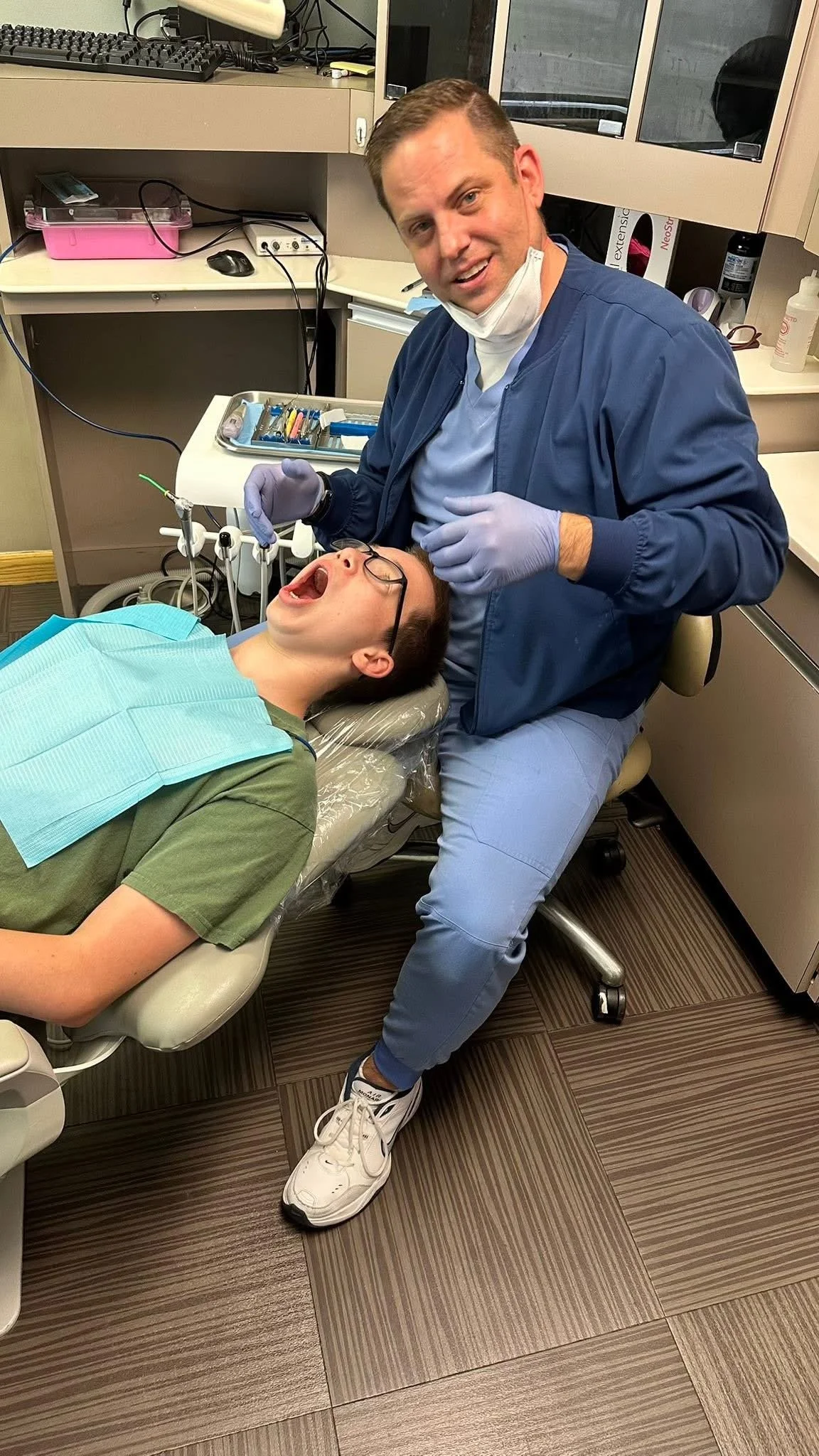 A dentist or dental assistant wearing a blue coat, gloves, and a mask, leaning over a patient lying in a dental chair, about to perform a dental procedure. The patient has their mouth open and is wearing glasses, with a dental bib on.