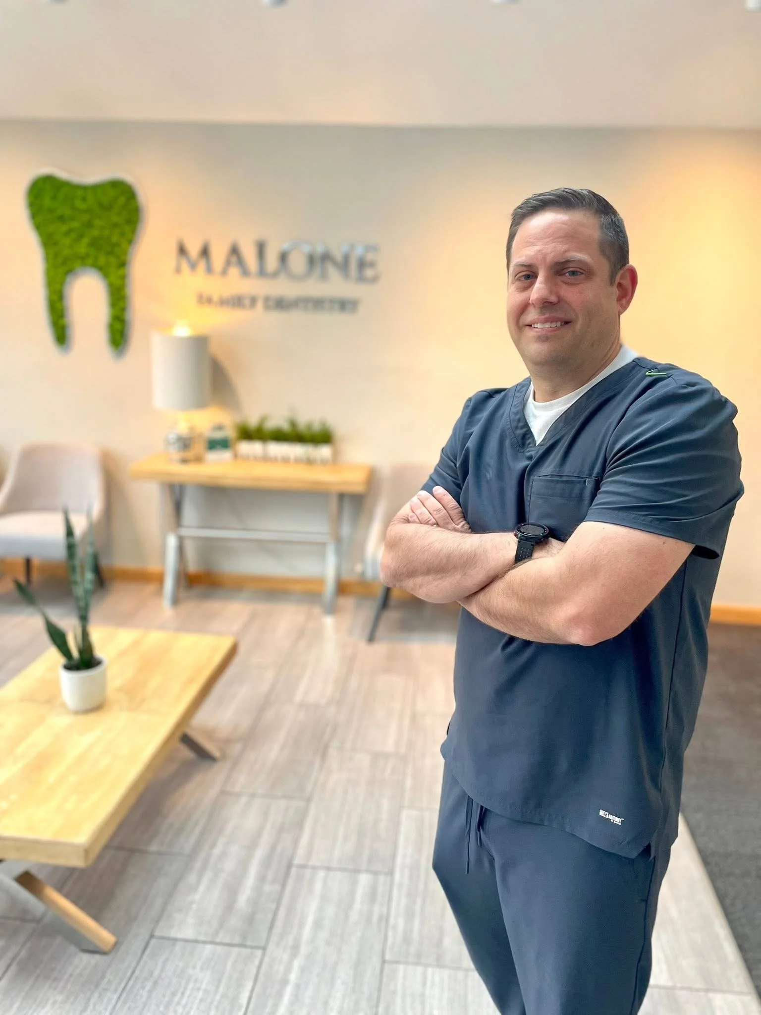 Male dentist standing with arms crossed inside dental office, with a wall sign reading 'Malone Elevated Dentistry' and a tooth-shaped decor