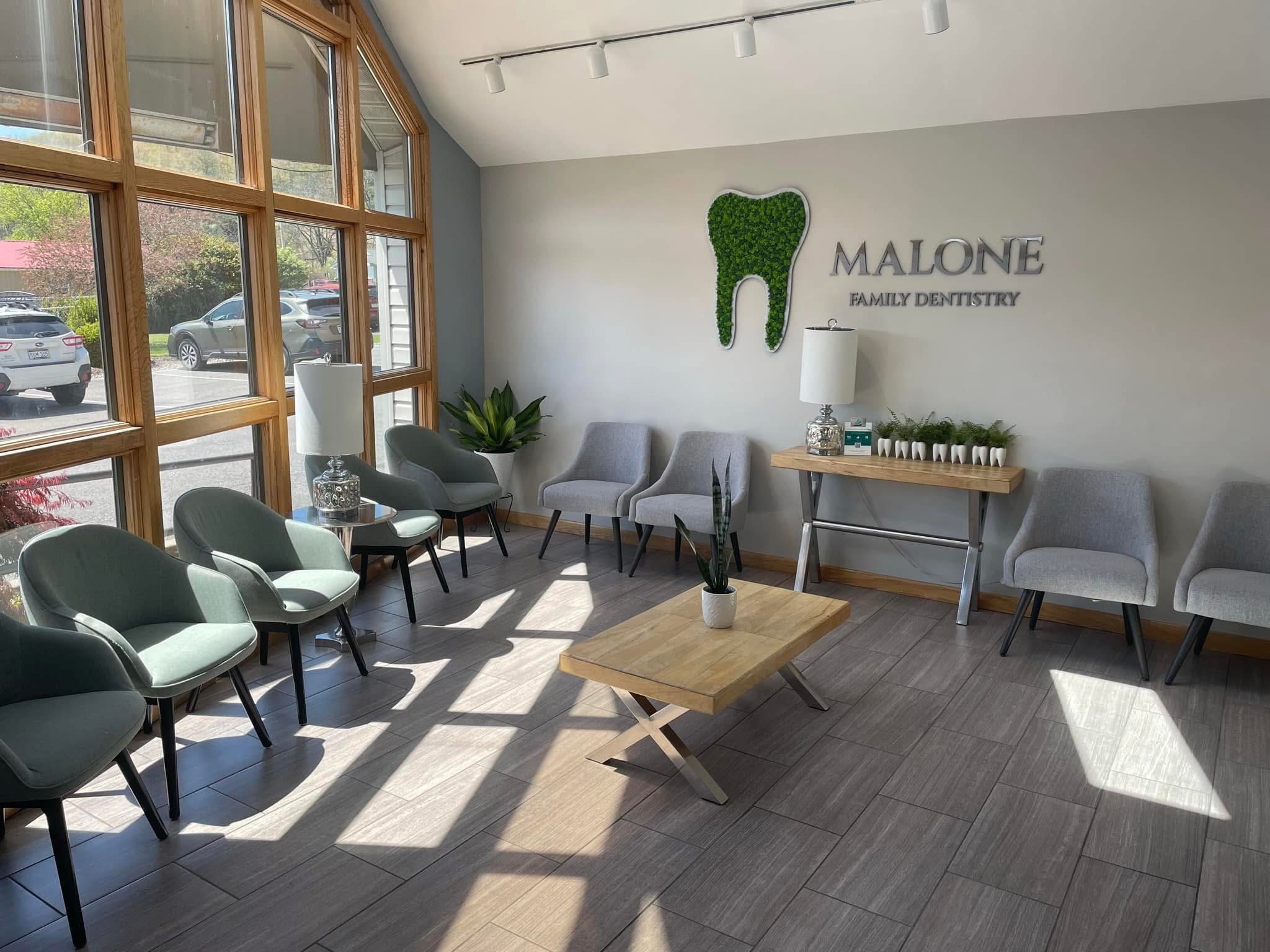 Waiting room at Malone Family Dentistry with light gray chairs, wooden tables, and a decorative green tooth on the wall, near large front windows letting in natural light.