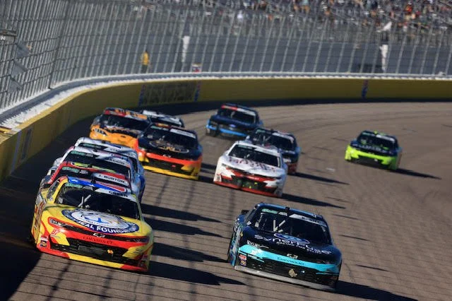 Multiple stock cars racing on a dirt track, with some cars near the front and spectators in the stands in the background.