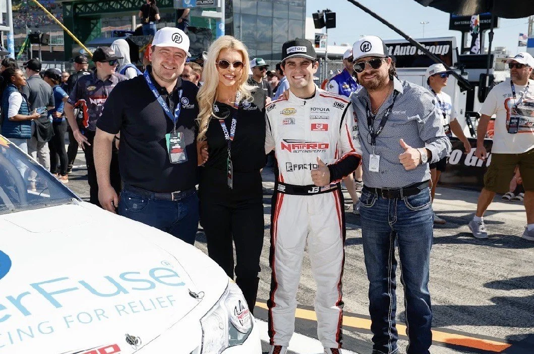 Four people standing in a racing event, with a race car partially visible in the foreground. The person second from the right is in a white racing suit, giving a thumbs-up, and everyone is smiling. There are numerous spectators and event staff in the