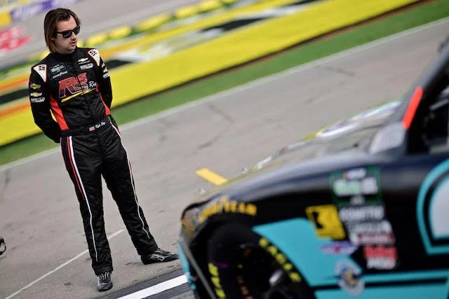 Kyle Sieg, male racing driver in a black racing suit with red and yellow accents, standing on a racetrack next to a race car, wearing sunglasses.