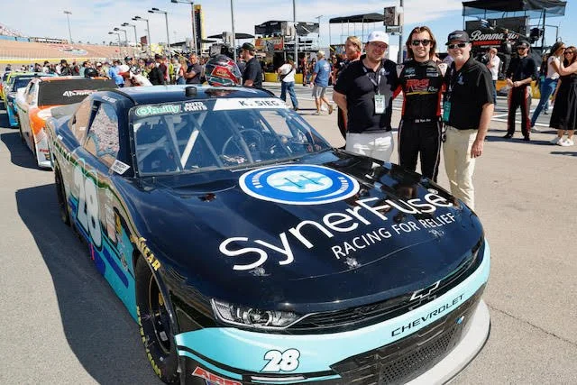 Three people standing next to a black and turquoise race car with the number 28 and Chevy logo, in a race track area with other cars and spectators in the background.