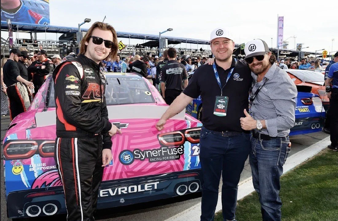 Three men standing in front of a pink race car at a racing event, with lots of people and cars in the background. The man on the left is wearing a racing suit and sunglasses, and the other two men are wearing casual clothes and sunglasses.