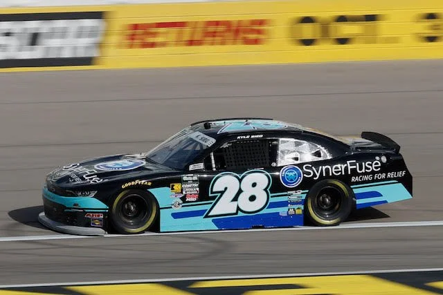 A black and blue NASCAR race car with the number 28 on the side, racing on a track. The car has sponsor logos including SynerFuse and Goodyear, with a blurred yellow and black background.