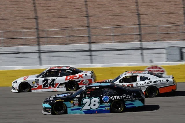 Three stock cars racing on a track with a brick wall and safety barriers in the background. The cars are numbered 24, 20, and 28, with various sponsor logos.