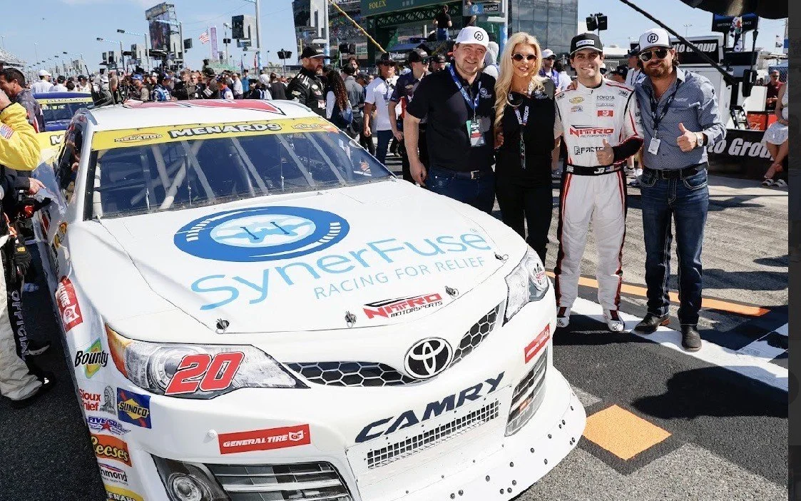 Group of people standing next to a race car at a racing event, with spectators in the background.