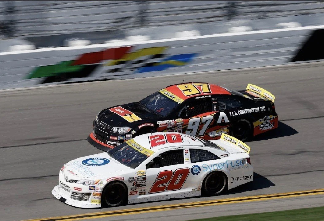 Two race cars on a track, one white with the number 20 and the other black with red and the number 97, racing side by side.