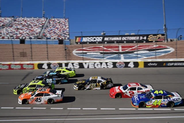 Race cars speeding on a NASCAR track at Las Vegas Motor Speedway with a large NASCAR banner and a grandstand in the background.