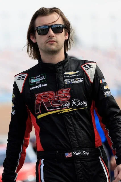 Race car driver, Kyle Sieg, in a black racing suit with red, white, and blue accents, wearing sunglasses, with a cloudy sky background.