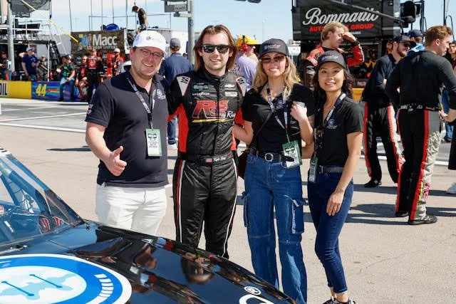 Kyle Sieg and others standing together at a racing event, with a race car partially visible in the foreground and a busy pit lane in the background.