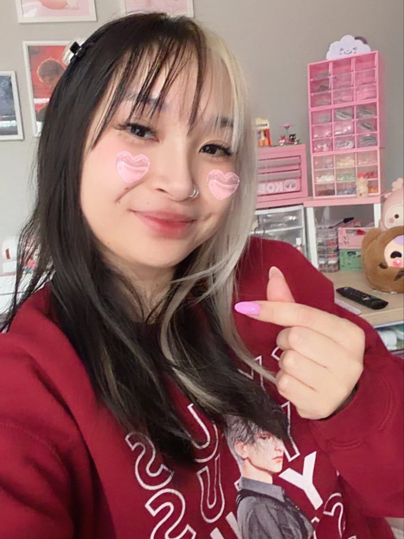 A young woman with multi-colored hair and pink painted nails taking a selfie in a room decorated with pink storage units, plush toys, and framed pictures on the wall.