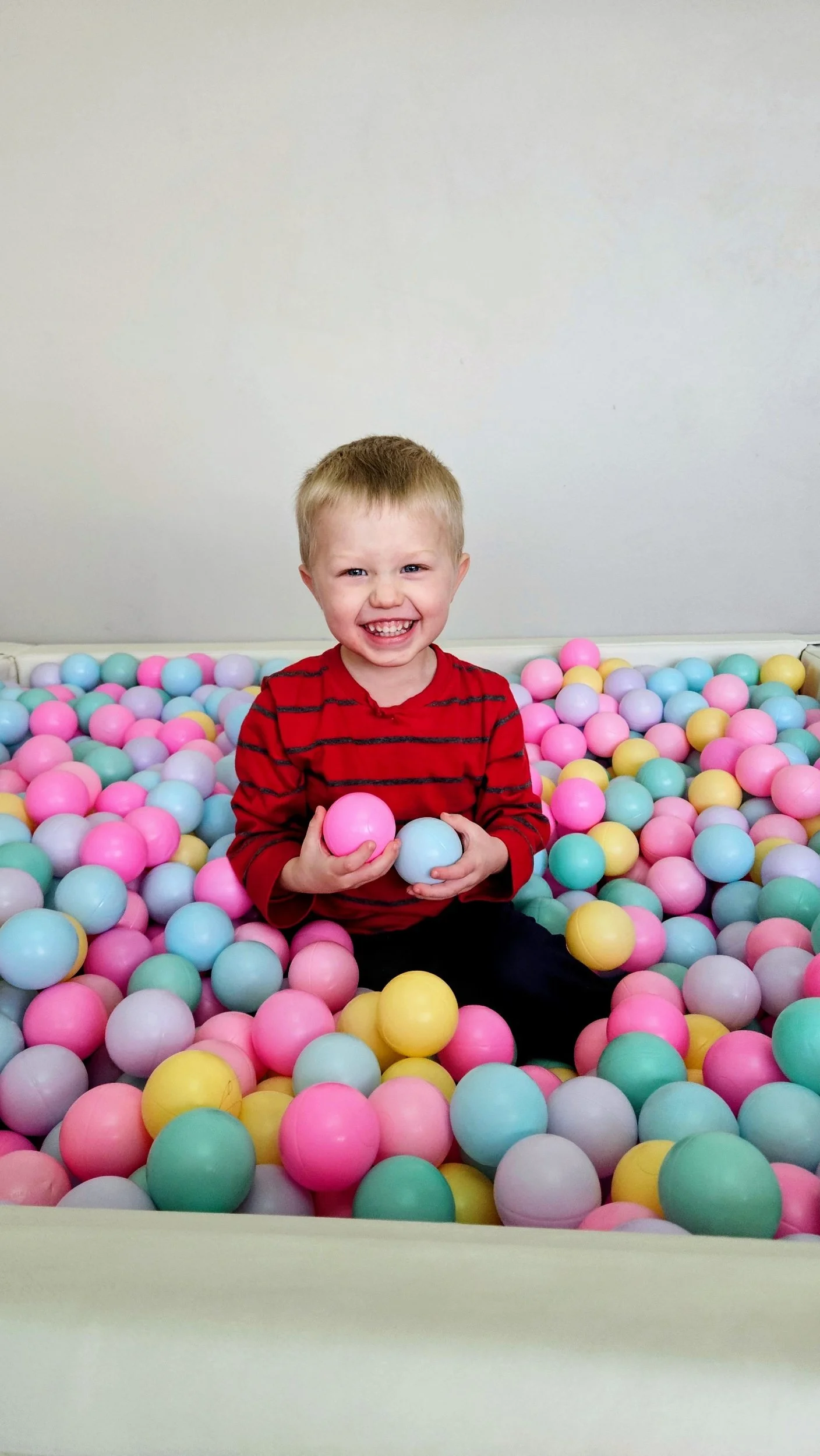A little boy sitting in a ball pit, smiling, and holding two balls