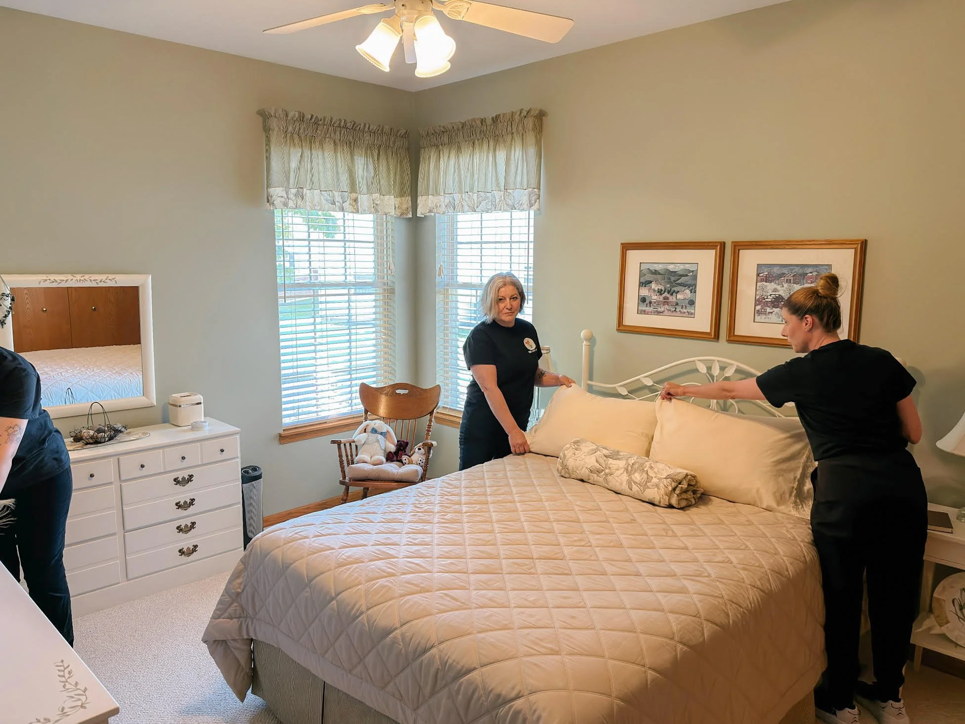 A bedroom with a large bed and beige quilt, two women making the bed, a window with striped curtains, a framed wall art, a white dresser with decorative items, and a ceiling fan with lights.