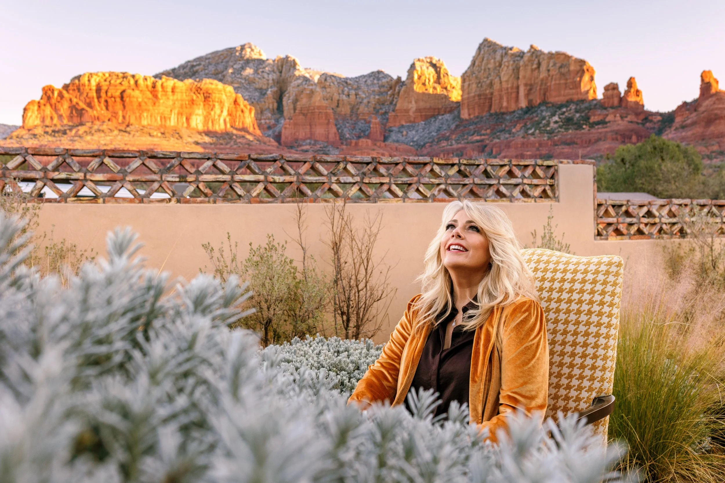 A woman with blonde hair smiling and looking upward, sitting outdoors with a desert mountain landscape in the background, during sunset or sunrise.