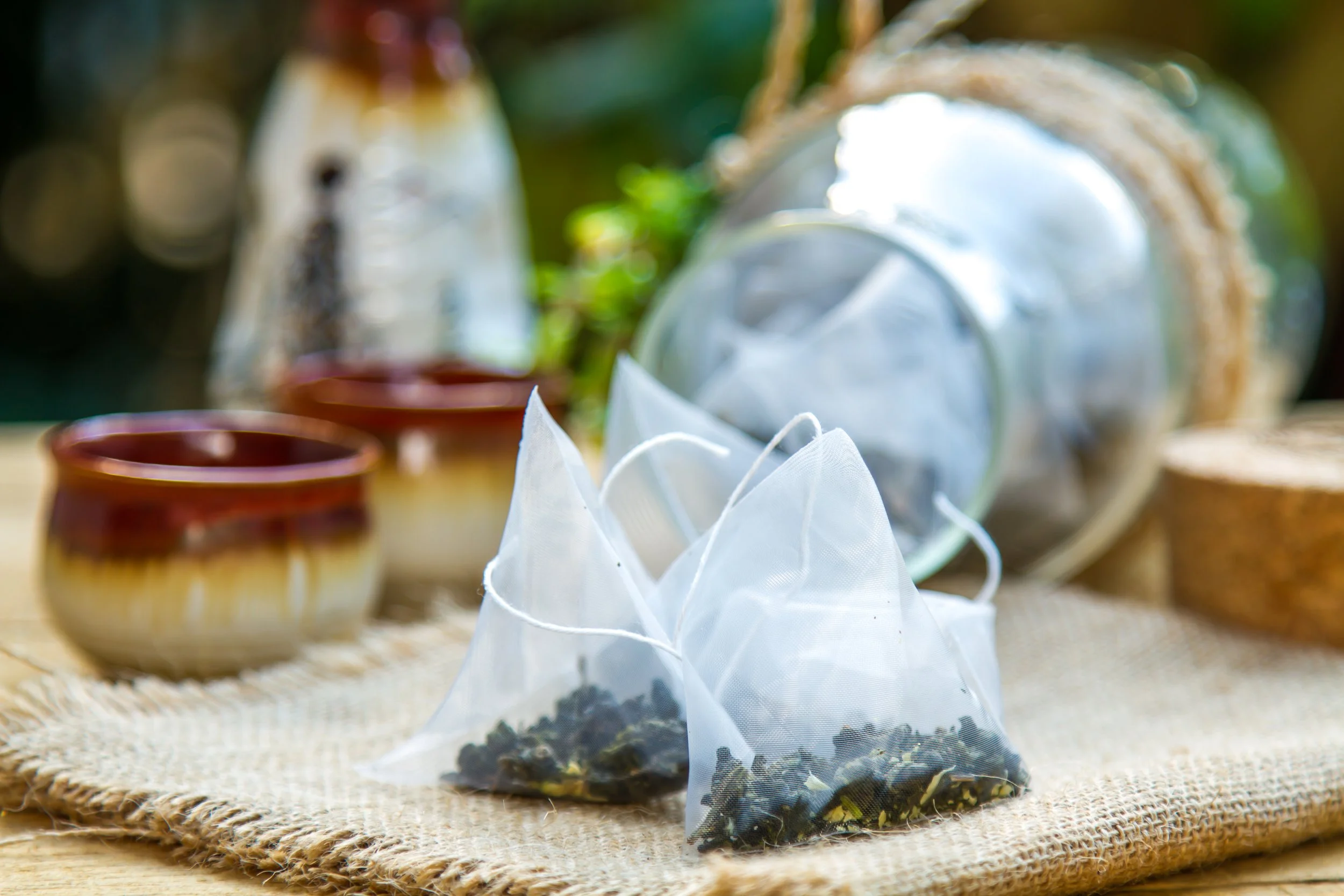Small transparent bag with dried tea leaves on a woven mat, with two small empty bowls and a white fabric bag in the background.