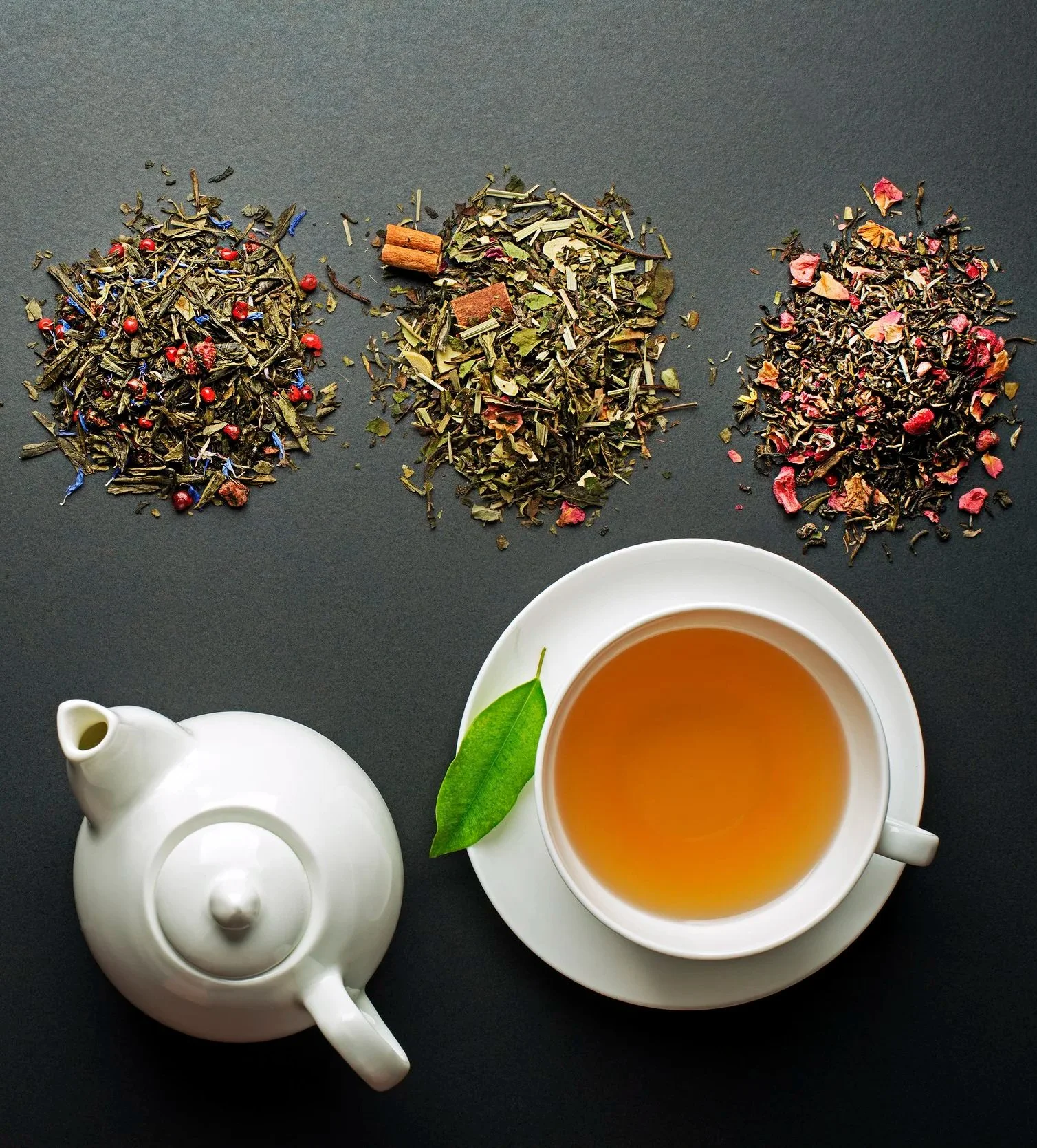 Three piles of loose herbal tea leaves on a dark surface, with a white teapot and a cup of brewed tea placed on a saucer, accompanied by a green leaf.