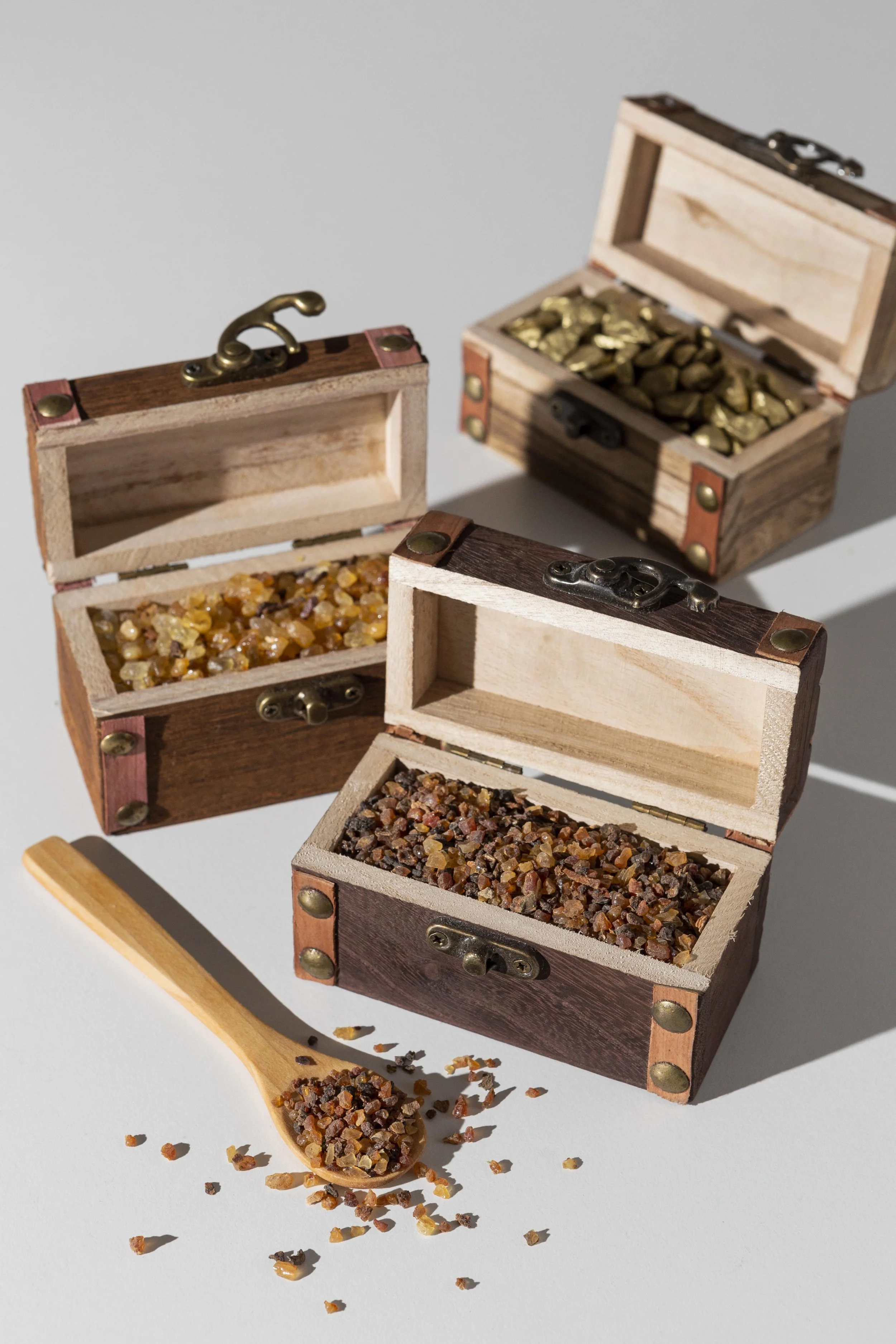 Several small wooden boxes filled with different types of colored sand or small rocks, some open with loose material spilling out, and a wooden spoon with loose material on it, all on a gray background.