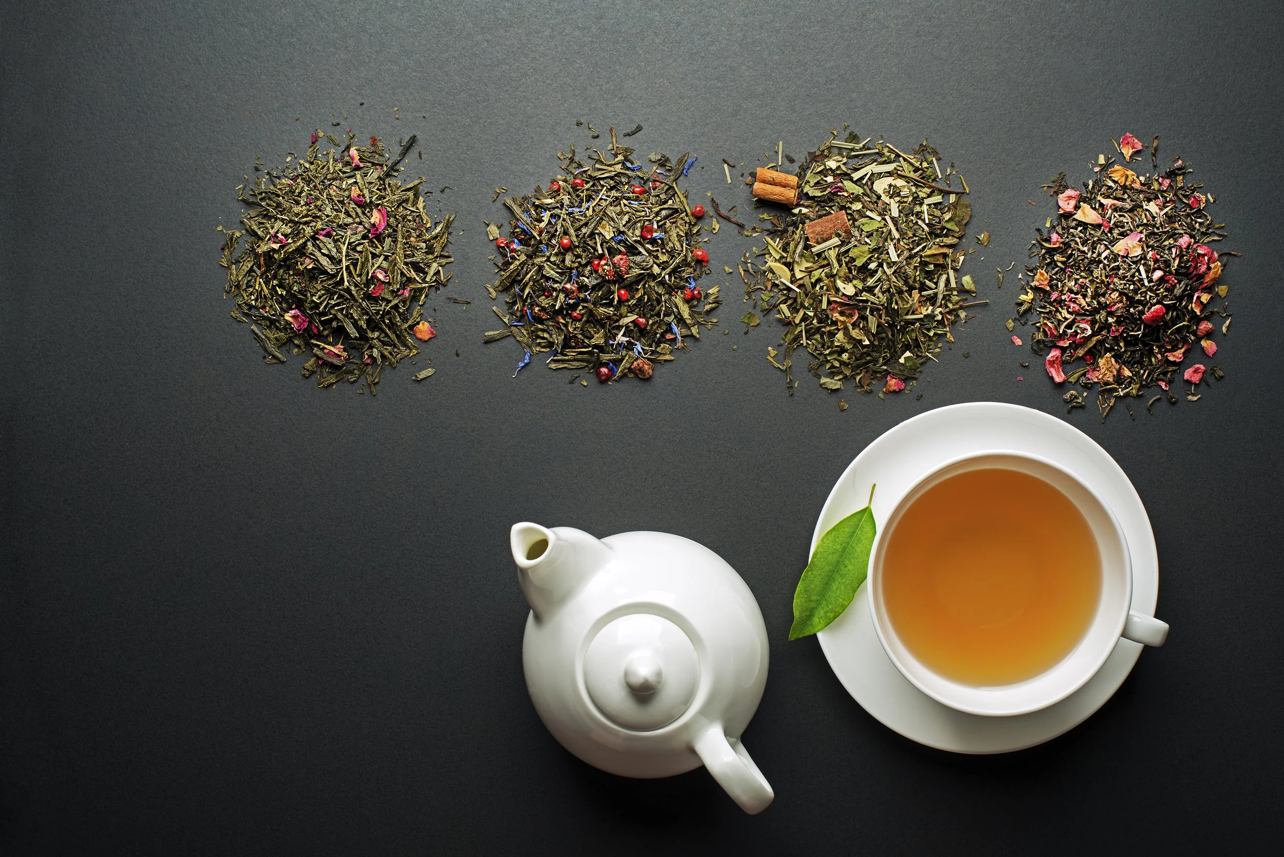 Loose dried herbs, spices, and flower petals arranged on a dark surface, alongside a white teapot and a cup of hot tea with a green leaf.
