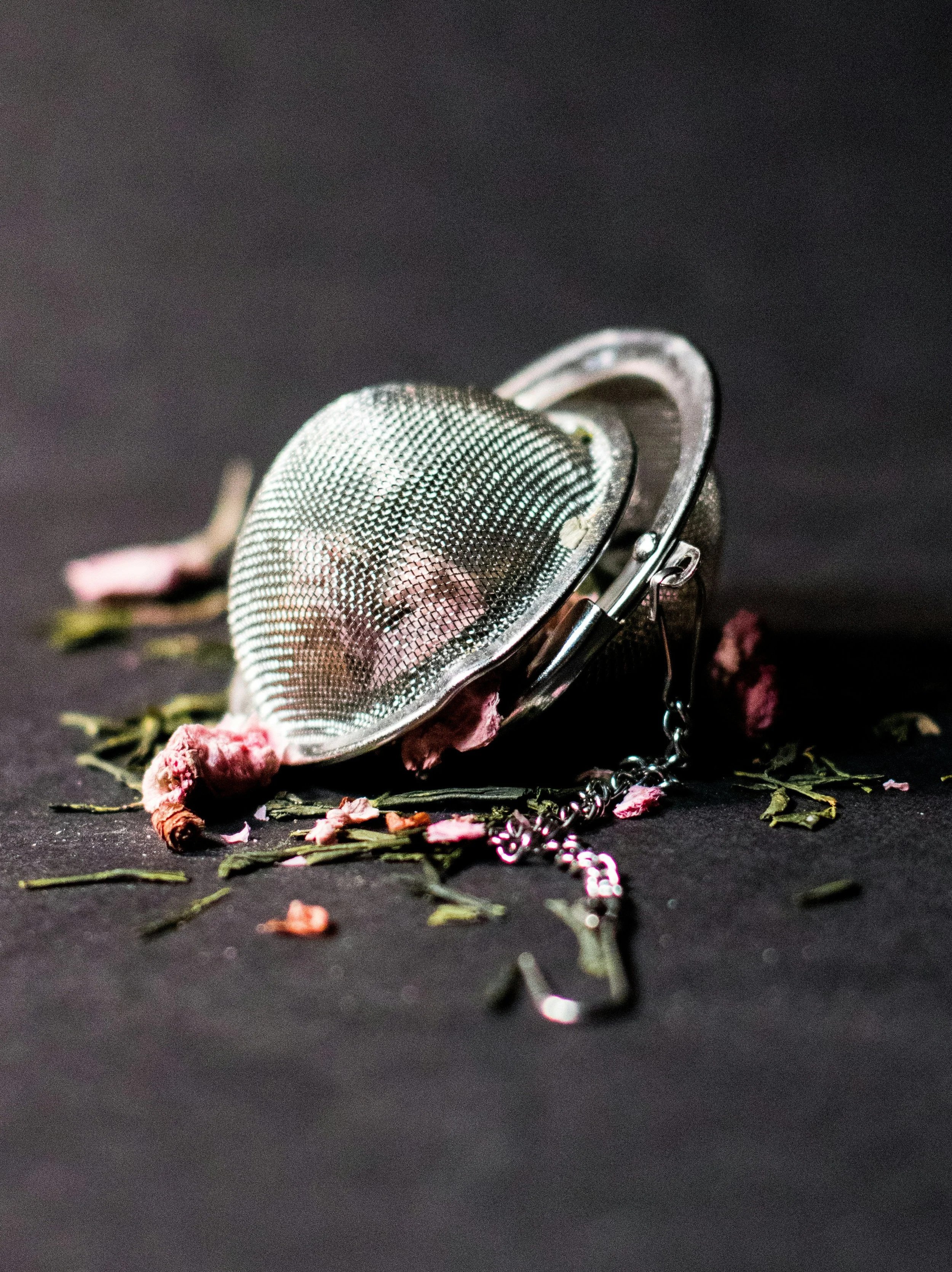 A close-up of a metal mesh sieve with a chain attached, surrounded by scattered dried flower petals and green leaves on a dark surface.