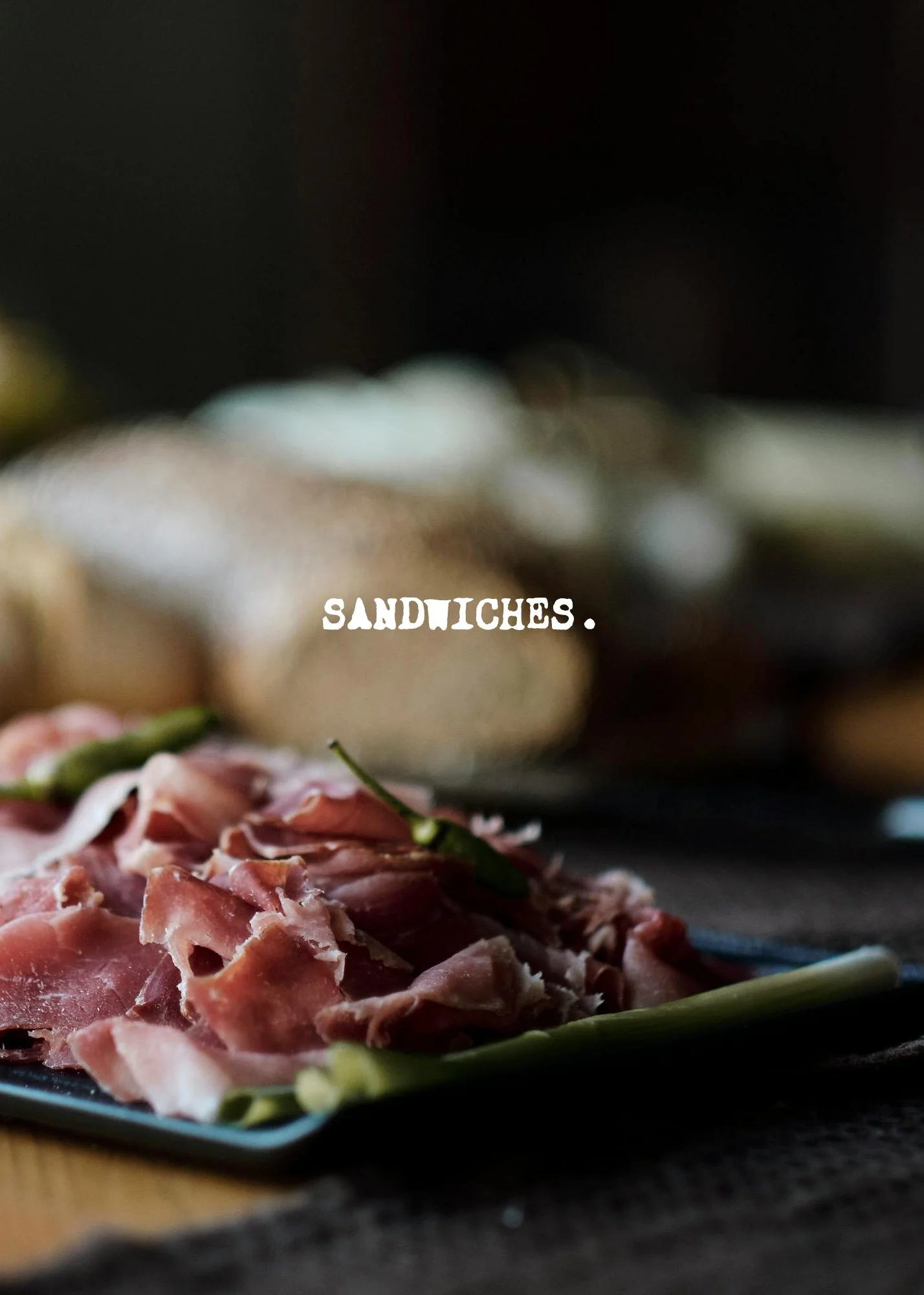 Close-up of a black platter with sliced cured ham, green beans, and a background of blurred dark area with some bread in the distance. The word 'SANDWICHES.' is written in white text at the center of the image.