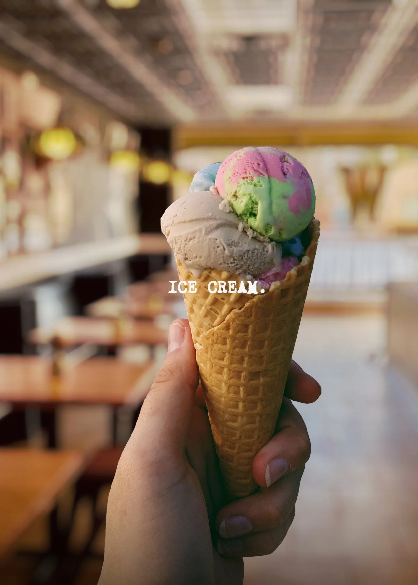 Close-up of a hand holding a waffle cone with three scoops of colorful ice cream with a blurred indoor restaurant background.