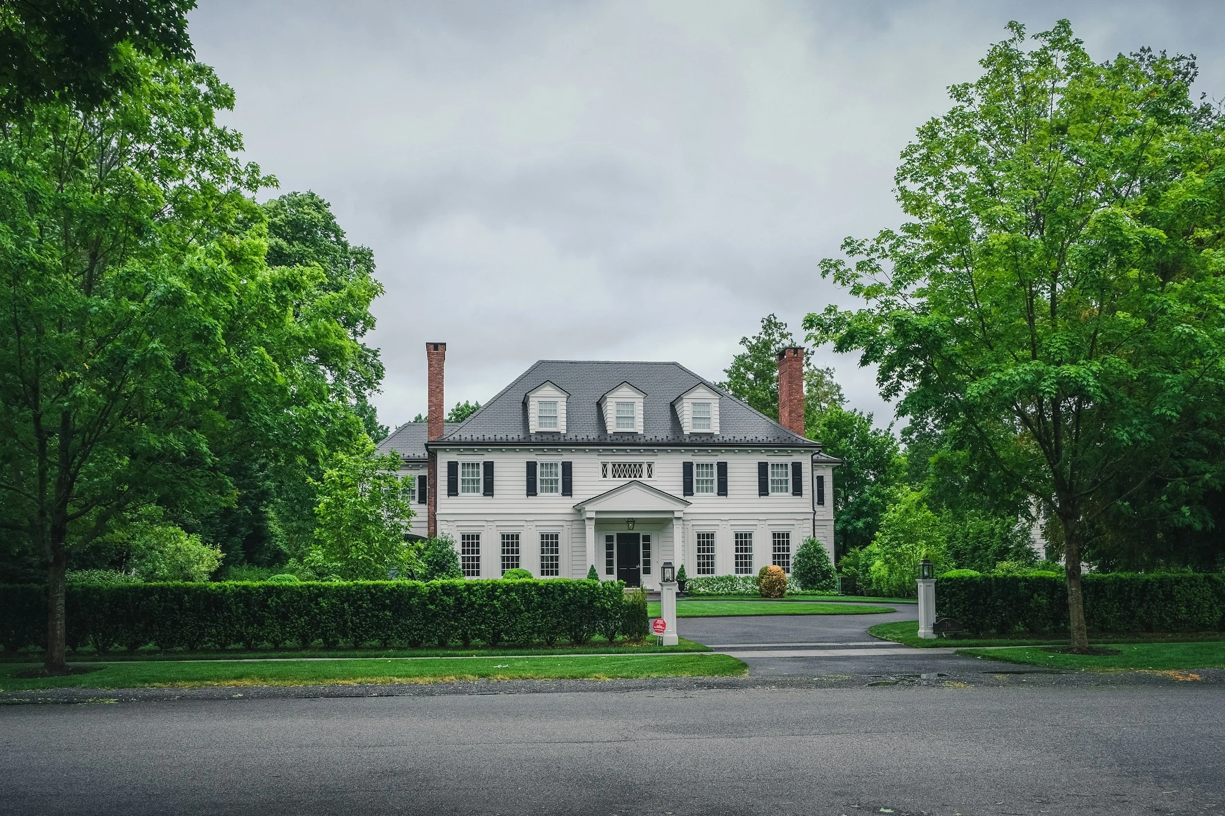 A large, white, two-story house with black shutters, a dark gray roof, and two brick chimneys, surrounded by green trees and a well-maintained lawn.
