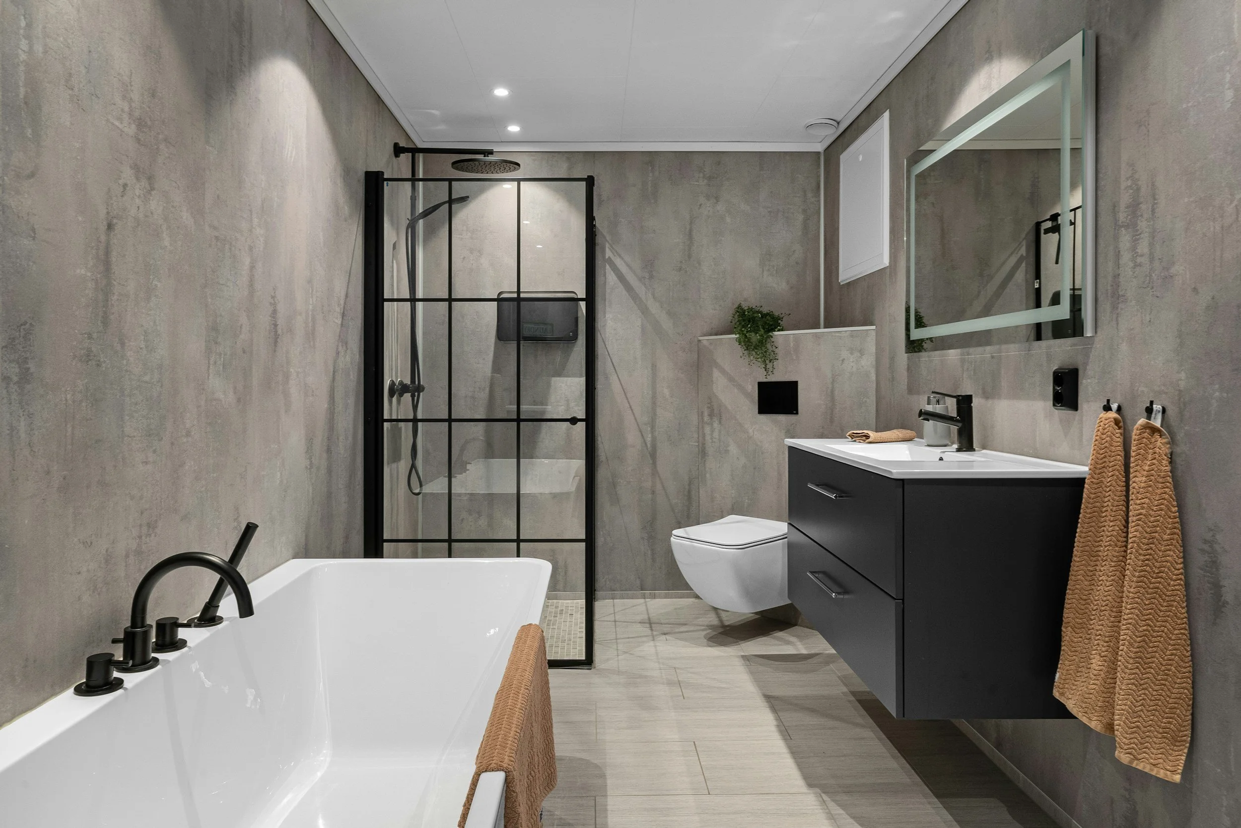 Modern bathroom with a white bathtub, a glass-enclosed shower, a wall-mounted toilet, and a black vanity with a mirror, beige towels, and wall decor.