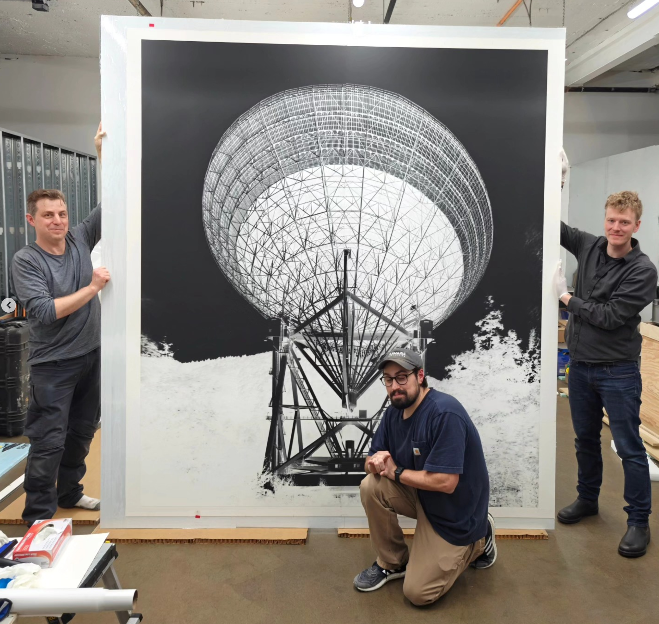 Three men pose with a large black and white photograph of a radio telescope, with two standing on either side and one kneeling in front.