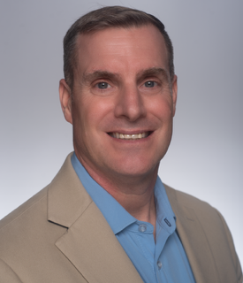 Portrait of a smiling man with short brown hair, wearing a beige blazer and a blue dress shirt against a neutral background.