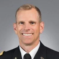 Portrait of a man in a military uniform smiling against a gray background.