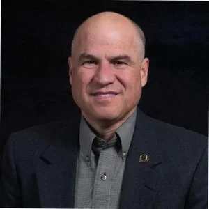 A middle-aged man with a bald head wearing a dark suit and a gray shirt, smiling at the camera against a dark background.